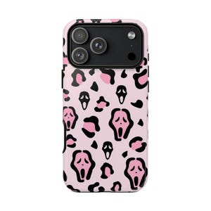 Può includere: Una custodia per smartphone con un motivo ripetuto di icone di maschere fantasma nere e rosa, intervallate da macchie leopardate nere e rosa su uno sfondo rosa chiaro. La custodia presenta un ritaglio nero per il modulo fotocamera in alto.