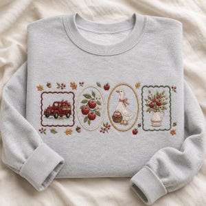 Könnte beinhalten: Ein hellgraues Sweatshirt mit Rundhalsausschnitt und vier gestickten rechteckigen und ovalen Rahmen. Die Motive zeigen einen roten Lastwagen mit Äpfeln, einen Apfelzweig, eine weiße Gans mit einem Korb und eine Vase mit Blumen.