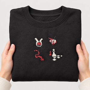 May include: A black crewneck sweatshirt featuring four small embroidered designs on the chest: a pair of hands holding a red apple, a red and white butterfly, a red snake, and a white chess queen piece with a red pawn on a checkered board.