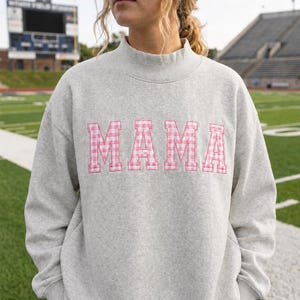 May include: A light gray heathered sweatshirt with a mock neck collar. The word MAMA is embroidered across the chest in a pink and white gingham check pattern with a thin pink outline surrounding each block letter of the text.