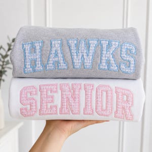 Op de afbeelding: Een grijs sweatshirt met het woord HAWKS in blauwe ruitjes-blokletters en een wit sweatshirt met het woord SENIOR in roze ruitjes-blokletters. Beide kledingstukken zijn voorzien van grote, gestikte letters in varsity-stijl.