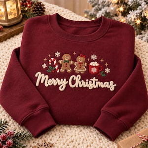 May include: A deep burgundy crewneck sweatshirt featuring an embroidered design of two gingerbread figures, a candy cane, and a mug of hot cocoa with marshmallows. The text Merry Christmas is embroidered in white script below the figures.