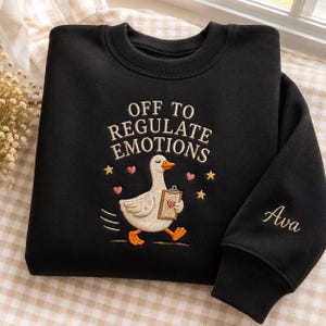 Puede incluir: Una sudadera negra de cuello redondo con texto bordado que dice OFF TO REGULATE EMOTIONS sobre un ganso blanco que sostiene un portapapeles con un corazón. El diseño incluye pequeños corazones rosas y estrellas amarillas. El nombre Ava está bordado en la manga.