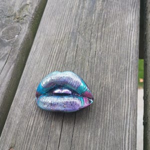 May include: A sculpted object shaped like a pair of lips with a metallic finish. The surface features a blend of iridescent blue, purple, and teal colors with a glossy, reflective texture that highlights the contours of the upper and lower lips.