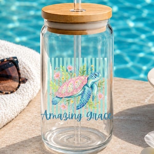 May include: A clear glass tumbler with a bamboo lid and a clear glass straw. The glass features a graphic of a sea turtle surrounded by pink and yellow flowers against a light blue striped background, with the text Amazing Grace printed below.