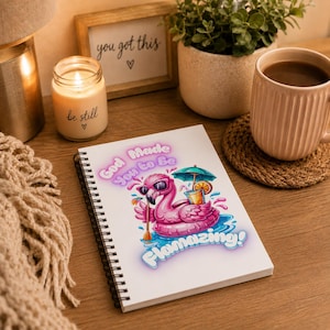 May include: A spiral-bound notebook featuring a graphic of a pink flamingo floatie wearing sunglasses, holding a drink with an umbrella, and a paddle. The text reads God Made You to be Flamazing! in purple and blue bubble letters on a white cover.