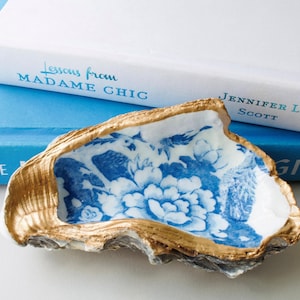 May include: An oyster shell trinket dish featuring a white interior with a blue floral pattern and a metallic gold painted rim. The exterior of the shell has a natural textured grey finish, contrasting with the decorative interior and gold edge.