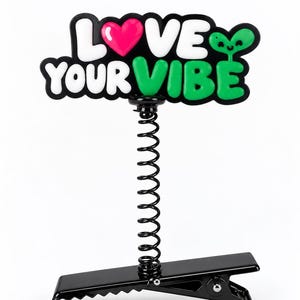 以下が含まれることがあります： LOVE YOUR VIBEと書かれた黒、白、緑、ピンクの看板が付いた黒いヘアクリップ。看板にはOの代わりにピンクのハートが描かれ、テキストの横には小さな緑色のエイリアンのキャラクターが配置されています。看板は垂直の金属製スプリングで支えられています。