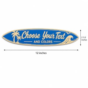 Puede incluir: Un letrero con forma de tabla de surf en azul con gráficos color crema, que presenta palmeras, una ola y el texto "Choose Your Text AND COLORS". El letrero mide 30,48 cm de largo y 6,35 cm de alto, según indican las flechas de medida.