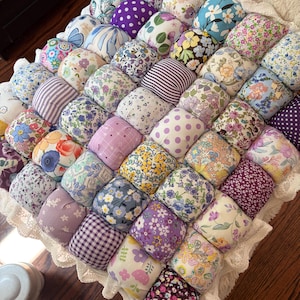 Pode incluir: Uma colcha quadrada de tipo puff construída com numerosos quadrados de tecido individuais que apresentam uma variedade de padrões florais, de bolinhas e de riscas em tons de roxo, azul, amarelo e rosa, com um rebordo de renda branca.