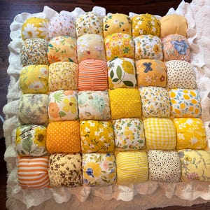 May include: A square patchwork quilt featuring a grid of thirty-six stuffed fabric squares in various shades of yellow, orange, and white. The squares display diverse floral, polka dot, and striped patterns, finished with a white lace trim border.