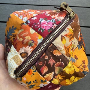 May include: A small, cube-shaped fabric pouch featuring a patchwork design of autumn-themed patterns including fall leaves, acorns, and floral motifs in shades of orange, brown, yellow, and red, secured with a dark brown zipper and metal pull.