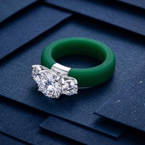 Op de afbeelding: Een ring met een dikke, effen groene band van glad materiaal, geaccentueerd door een zilverkleurige metalen zetting die drie ronde, heldere, gefacetteerde stenen in een horizontale rij boven op de band vasthoudt.