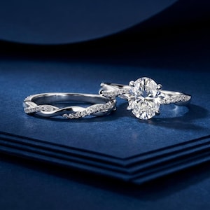 May include: A silver-toned engagement ring set featuring an oval-cut center stone and a matching band. Both rings have a twisted design accented with small, clear pavé-set stones along the bands, creating a cohesive and sparkling jewelry set.