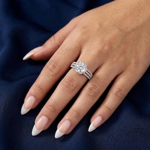 Puede incluir: Un anillo de tono plateado con una gran piedra central redonda de talla brillante rodeada por un halo de piedras más pequeñas. La banda se divide en tres filas distintas, cada una incrustada con pequeñas piedras brillantes, creando un aspecto ancho y superpuesto.