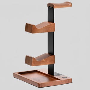 May include: A vertical headphone stand featuring a black metal central pillar and three horizontal wooden supports with curved cradles. The base is a rectangular wooden tray with a small circular button integrated into the right side of the surface.