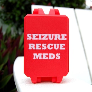 May include: A rectangular red plastic case with a textured surface and a hinged top. The words SEIZURE RESCUE MEDS are printed in bold white capital letters on the front center of the case, which features a secure latch mechanism at the top.
