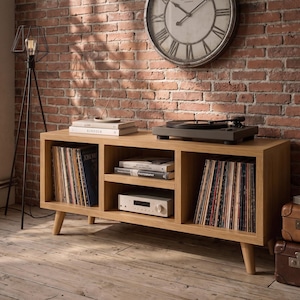 May include: A light oak media console with four tapered legs, featuring two large side compartments filled with vinyl records, a central shelf holding books and a silver audio receiver, and a black turntable placed on the top surface.