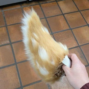 May include: A faux fur tail accessory featuring a blend of golden tan and white long-pile synthetic fibers. The tail has a tapered, curved shape and includes a white elastic band attachment at the base for securing the item to a belt or clothing.