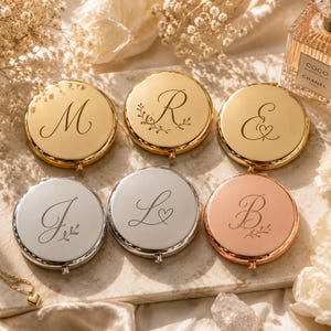 May include: Six round compact mirrors in gold, silver, and rose gold finishes. Each mirror features a unique engraved letter or symbol, including M, R, &, J, L, and B, with some designs incorporating delicate leaf or heart motifs on the metal surface.