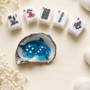 May include: Five white rectangular mahjong tiles with black, red, and green symbols and numbers, arranged in a semi-circle above an oyster shell dish containing two translucent blue dice and a small blue dolphin figurine.
