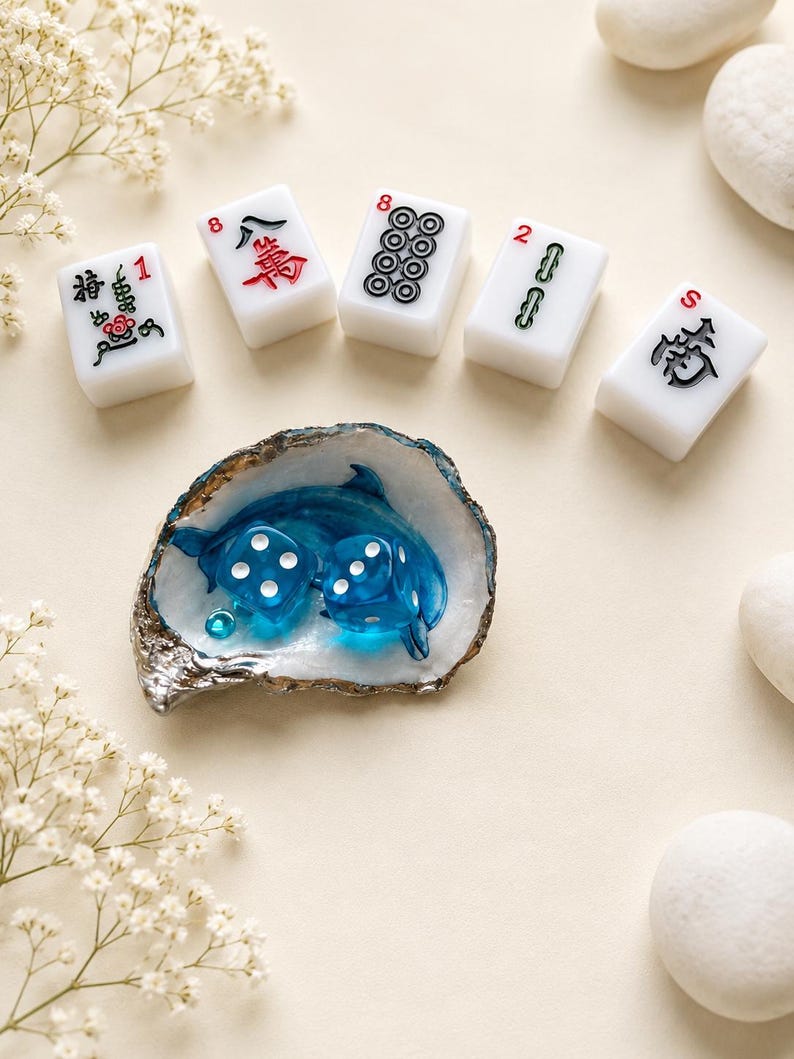 May include: Five white rectangular mahjong tiles with black, red, and green symbols and numbers, arranged in a semi-circle above an oyster shell dish containing two translucent blue dice and a small blue dolphin figurine.