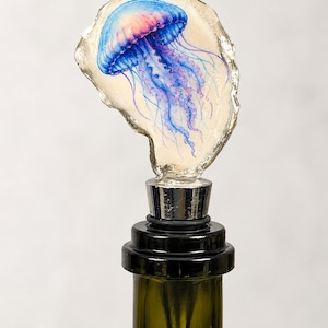 May include: A bottle stopper featuring a hand-painted jellyfish on a natural oyster shell. The jellyfish displays vibrant blue and pink hues with long, flowing tentacles. The shell is mounted on a silver-toned metal base with a black rubber seal.