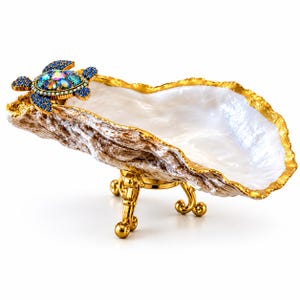 May include: A decorative oyster shell dish featuring a gold-trimmed edge and a gold metal stand with three legs. A jewelled turtle ornament with blue and iridescent crystals is attached to one end of the shell, which has a white pearlescent interior.