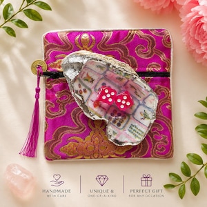 May include: A square magenta fabric pouch with a gold brocade pattern, a black zip, and a purple tassel attached to a gold coin. Resting on the pouch is an oyster shell with a silver-painted edge, containing two small pink dice with white dots.