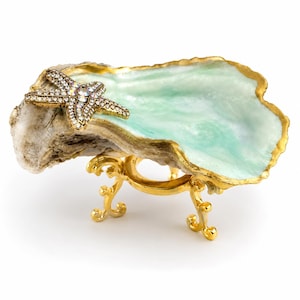May include: A decorative oyster shell dish featuring a light green interior, a gold-painted rim, and a gold-toned starfish accent encrusted with clear crystals. The shell is supported by a gold-colored metal stand with ornate, curved legs.