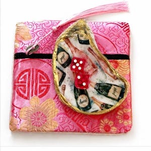 Peut inclure: Une pochette carrée en tissu rose avec un motif floral doré et un pompon rose, ornée d'une coquille d'huître décorative sur le devant. La coquille présente un bord doré et un motif de bouteilles de champagne avec deux dés rouges.