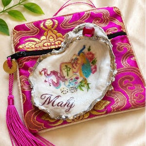 May include: A bright pink fabric pouch with gold brocade patterns, a black zip, and a pink tassel. Resting on top is a white oyster shell painted with a colourful dragon, mahjong tiles, a peacock, and the word Mahj, topped with a red gem.
