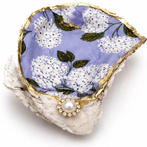 May include: A decorative oyster shell dish featuring a light purple interior with a white hydrangea floral pattern. The shell edge is painted with a metallic gold finish and is adorned with a single round white pearl surrounded by small clear crystals.