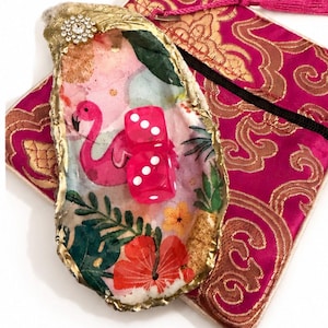 May include: A decorative oyster shell with gold-painted edges and a rhinestone accent, featuring a decoupage design of a pink flamingo, tropical leaves, and flowers. Two translucent hot pink dice rest on the shell, placed over a magenta fabric pouch.