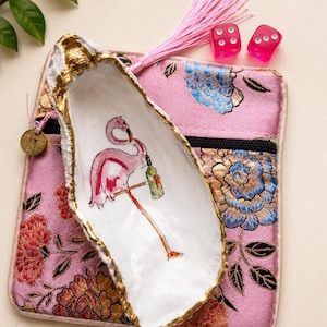 May include: A white oyster shell dish with a gold-painted rim, featuring a hand-painted pink flamingo holding a green bottle. It rests on a pink fabric pouch with a floral pattern, a light pink tassel, and two translucent pink dice.