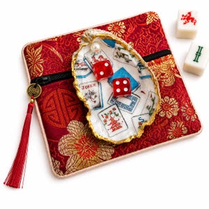 May include: A red fabric pouch with gold floral patterns, a black zipper, and a red tassel charm. Resting on top is a white oyster shell with gold-painted edges, featuring painted mahjong tiles, a pearl, and two red dice with white dots.