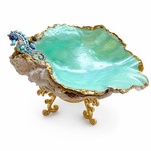 May include: A decorative bowl crafted from a natural shell with a shimmering mint green interior and a gold-painted rim. A blue and turquoise jeweled seahorse figure is attached to the edge. The bowl rests on three gold-colored metal legs.