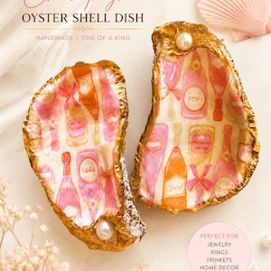 May include: Two oyster shell dishes with gold-gilded edges and a decoupage pattern of pink and orange champagne bottles. Each shell features a single white pearl accent. The text on the shells includes the word POP! and other labels.