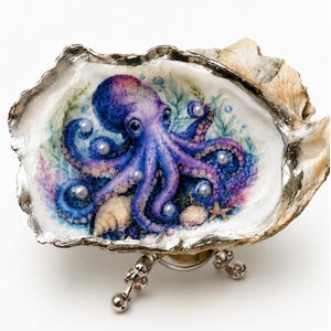 May include: A decorative oyster shell featuring a detailed illustration of a purple and blue octopus surrounded by green seaweed and small white pearls. The shell has a metallic silver-painted edge and rests on a small, three-legged silver metal stand.