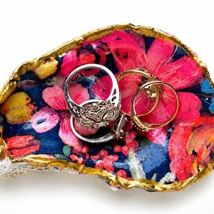 Puede incluir: Un plato decorativo para baratijas hecho de una concha de ostra con un patrón floral vibrante en tonos rosa, rojo y azul en el interior. Los bordes de la concha están pintados con un acabado dorado metálico. Tres anillos descansan dentro.