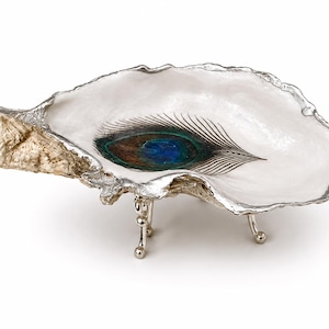 May include: A decorative oyster shell dish featuring a white interior with a central peacock feather design. The shell has a textured, natural exterior edge finished in silver and is supported by three small, polished silver metal legs.