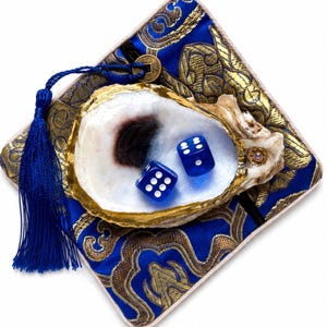 May include: A natural oyster shell with a gold-painted rim, containing two translucent blue dice with white pips. The shell rests on a square blue and gold patterned fabric cushion, which features a blue tassel and a small circular metal charm.