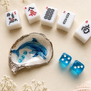 May include: A white oyster shell painted with a blue dolphin and three small blue beads, accompanied by five white mahjong tiles with black and red markings and two translucent blue dice with white pips, arranged on a light beige surface.