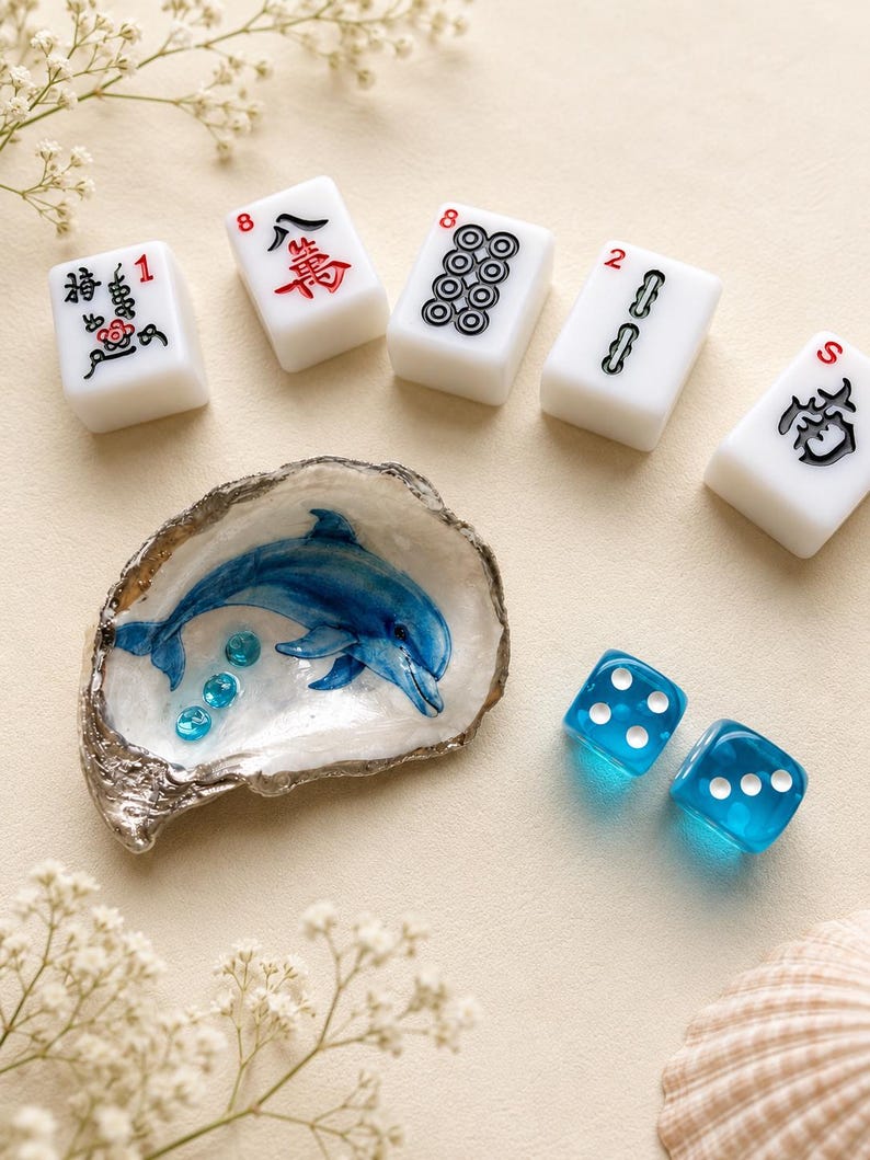 May include: A white oyster shell painted with a blue dolphin and three small blue beads, accompanied by five white mahjong tiles with black and red markings and two translucent blue dice with white pips, arranged on a light beige surface.