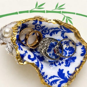May include: An oyster shell trinket dish featuring a white interior with a blue floral pattern and gold-painted edges. A single white pearl is attached to the shell rim. Three metal rings are resting inside the shell, including a gold band.