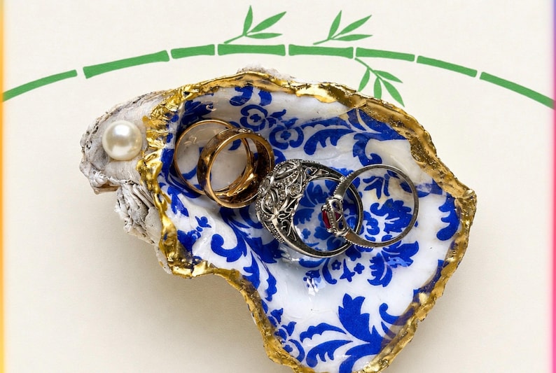 K&ouml;nnte beinhalten: Eine Austernschale als Schmuckschale mit wei&szlig;em Innenraum, blauem Blumenmuster und goldfarbenen R&auml;ndern. Eine einzelne wei&szlig;e Perle ist am Rand der Schale befestigt. Drei Metallringe liegen in der Schale, darunter ein goldener Ring.
