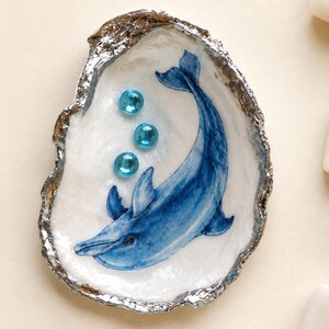 May include: A decorative oyster shell featuring a hand-painted blue dolphin illustration on the interior. The shell has a metallic silver-painted edge and is embellished with three small, round, translucent blue glass beads arranged vertically.