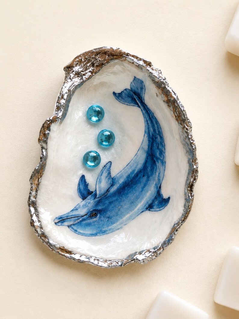 May include: A decorative oyster shell featuring a hand-painted blue dolphin illustration on the interior. The shell has a metallic silver-painted edge and is embellished with three small, round, translucent blue glass beads arranged vertically.