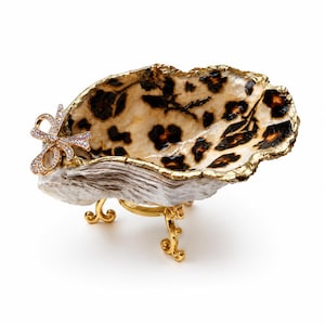 May include: A decorative oyster shell dish featuring a leopard print interior, a gold-painted scalloped rim, and a gold-toned metal stand with three curved legs. A small, crystal-encrusted bow ornament is attached to the side of the shell.