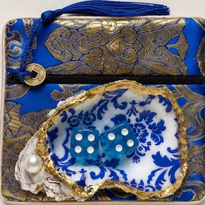 K&ouml;nnte beinhalten: Eine quadratische blau-goldene Brokattasche mit einer blauen Quaste und einem goldfarbenen M&uuml;nzanh&auml;nger am Rei&szlig;verschluss. Auf der Vorderseite befindet sich eine Austernschale mit blau-wei&szlig;em Blumenmuster, einer wei&szlig;en Perle und zwei blauen W&uuml;rfeln.